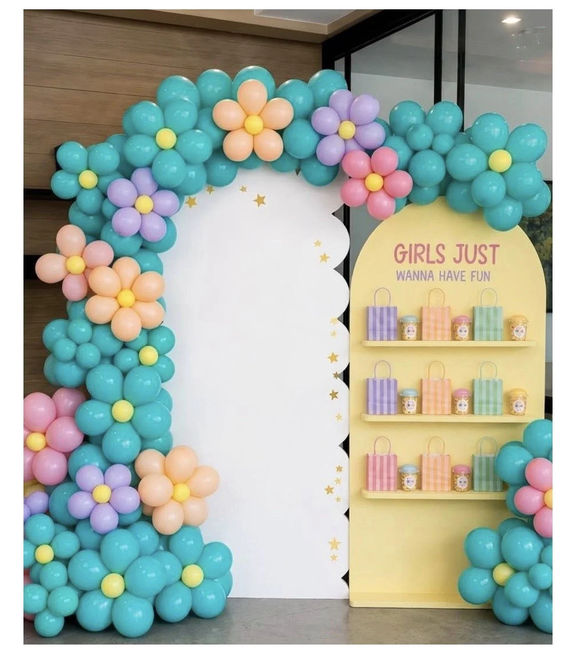 A teal balloon arch studded with peach, lilac and yellow balloon daisies beside a “girls just wanna have fun” favor wall