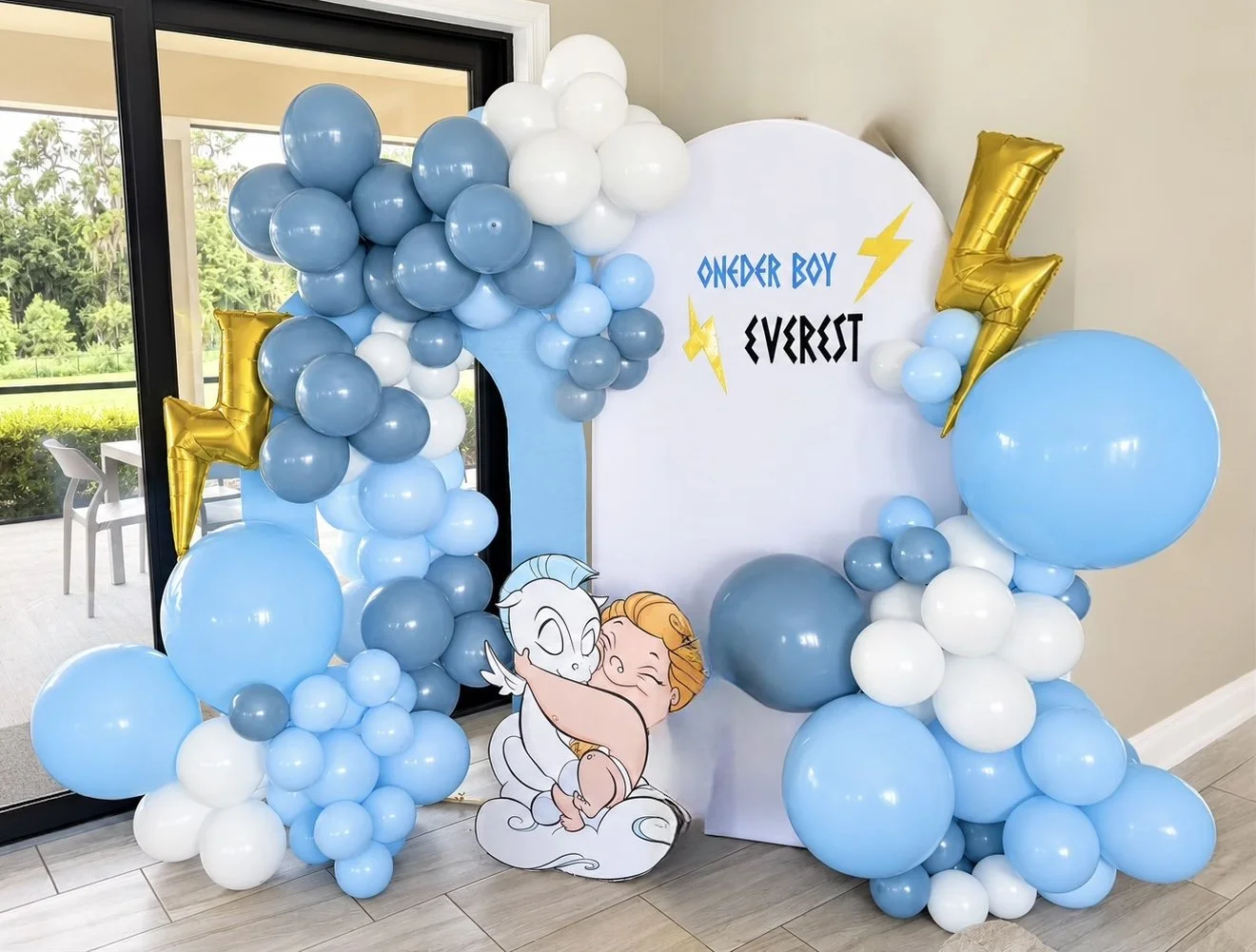 A dusty-blue, sky-blue and white balloon garland with gold lightning-bolt foils around a white arch reading “oneder boy Everest” with a baby Hercules cut-out