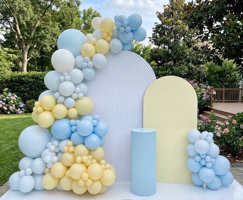 A pastel garland of powder-blue, butter-yellow and white balloons draped over a white ribbed arch, a pale-yellow panel and a blue pedestal in a garden