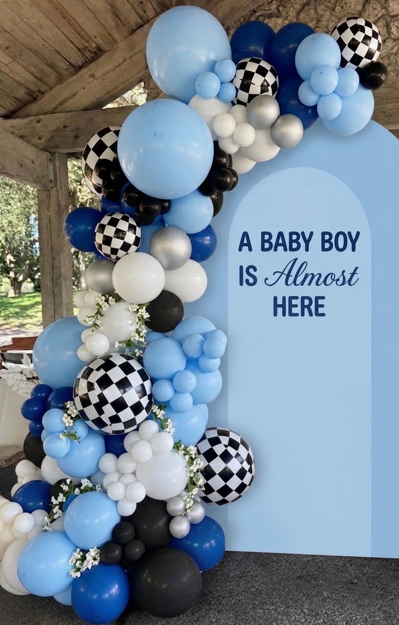 A blue, navy, black and white balloon garland with checkered-flag balloons and sprigs of white blossom beside a sky-blue arch reading “a baby boy is almost here”