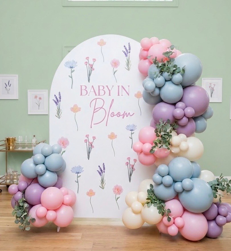 Pink, lilac, dusty-blue and cream balloon garlands with eucalyptus either side of a white arch printed with wildflowers reading “baby in bloom”