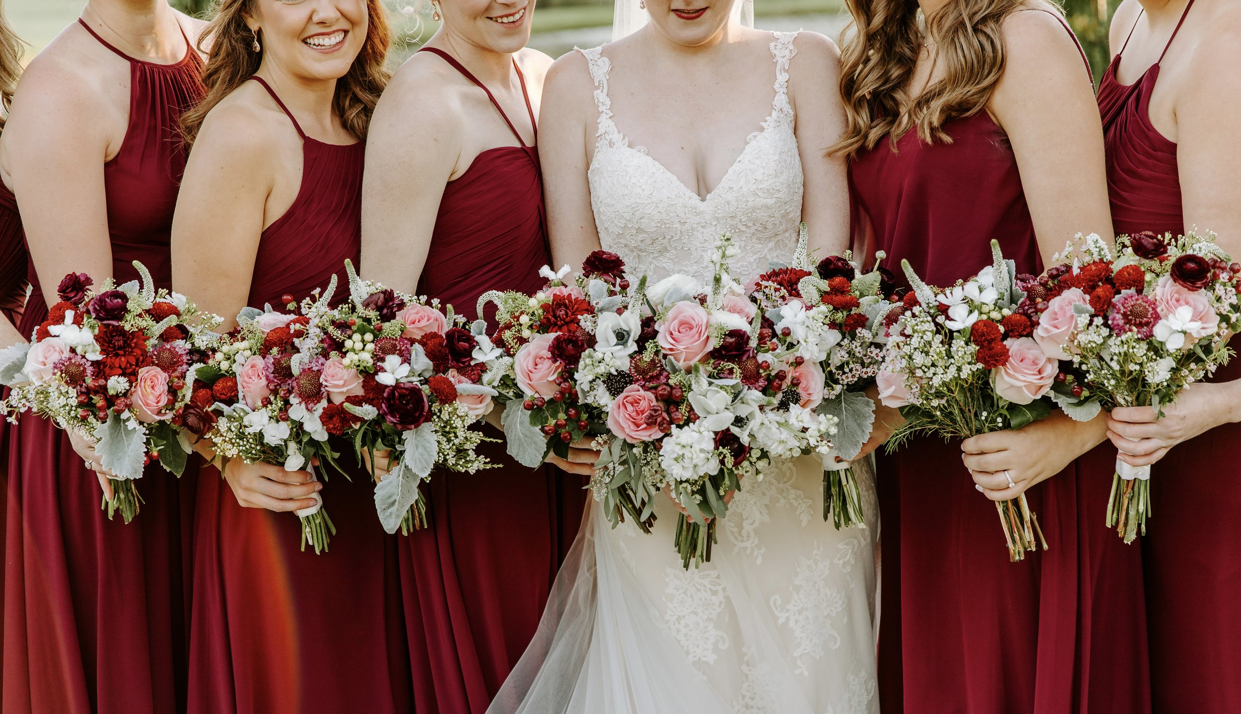 The bride and her bridesmaids in burgundy gowns holding bouquets of blush, coral and burgundy blooms