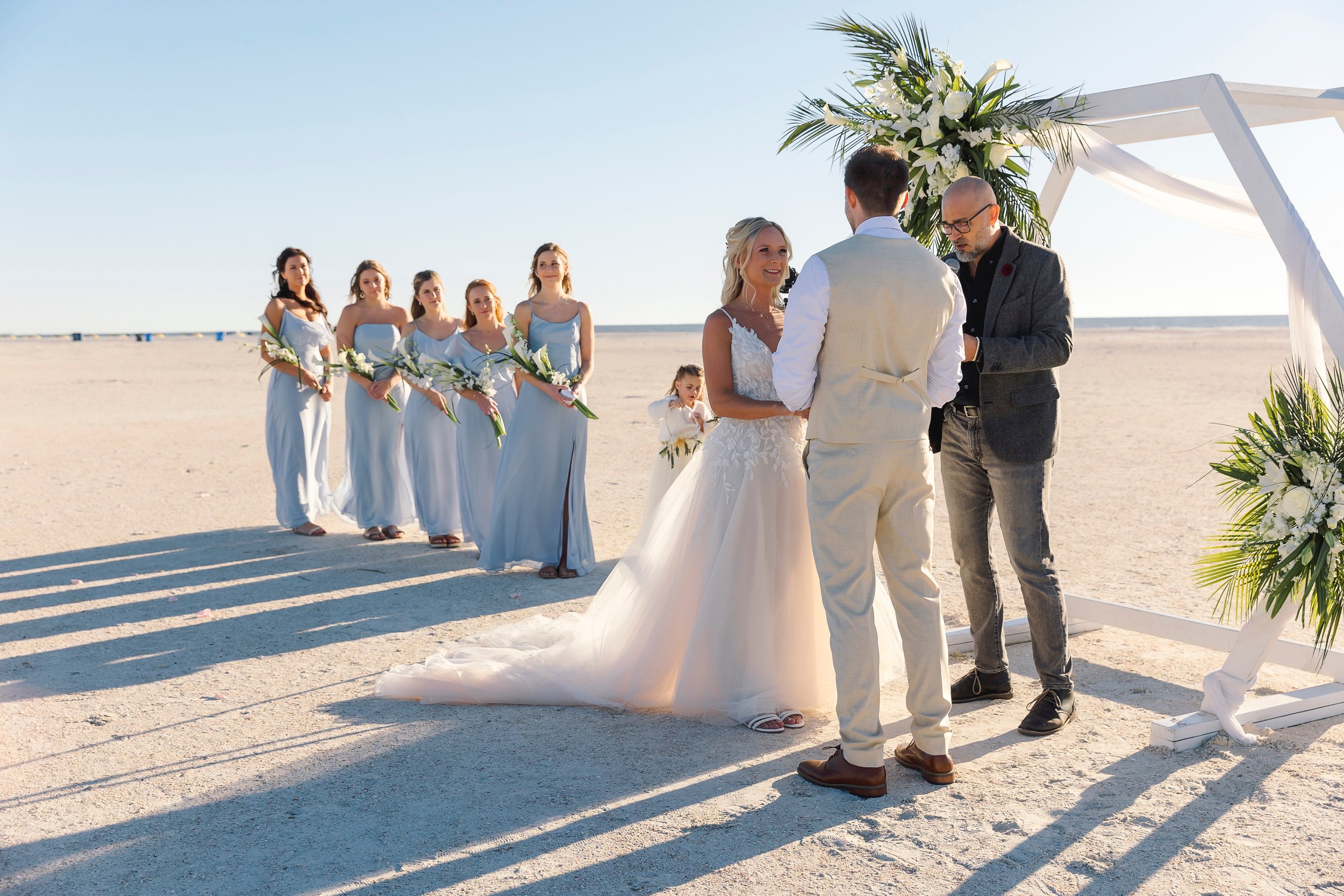 Bridesmaids in pale blue watch a beach ceremony at a white hexagon arch dressed with white florals and palm