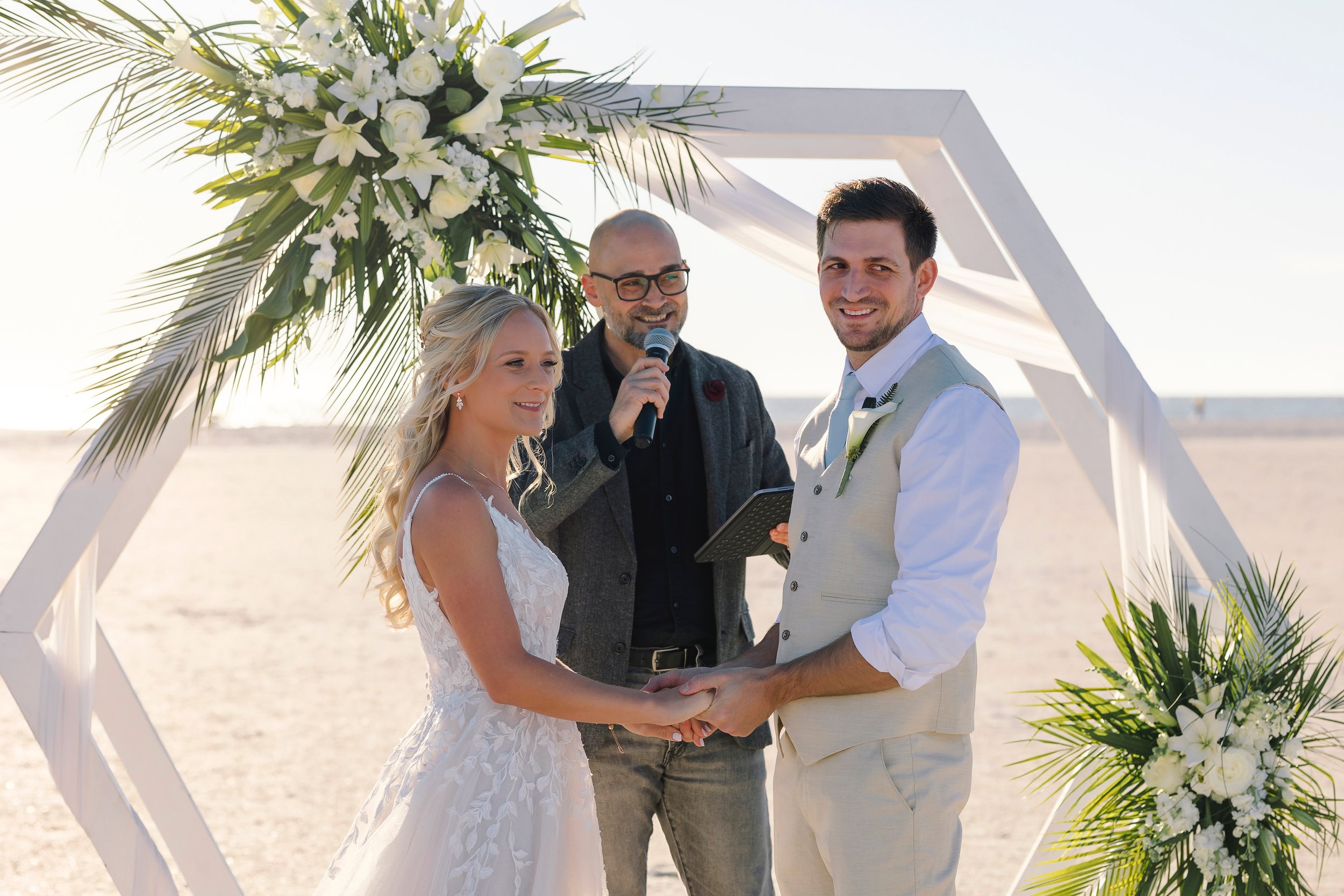 A couple exchange vows on the sand beneath a white hexagonal arch dressed with Julie's lilies, roses and palm fronds
