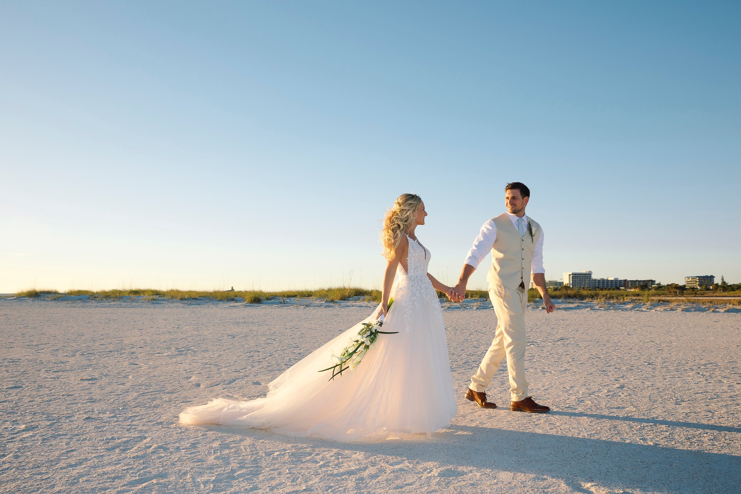 A bride and groom walk the sand at golden hour, her cascade bouquet of calla lilies trailing from one hand