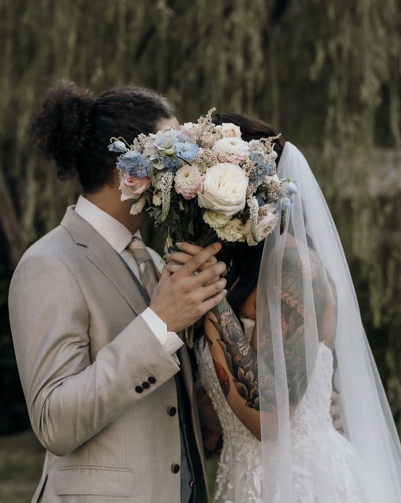 A groom in a sand-coloured suit holds his bride close, her bouquet of blush and blue blooms between them beneath a willow