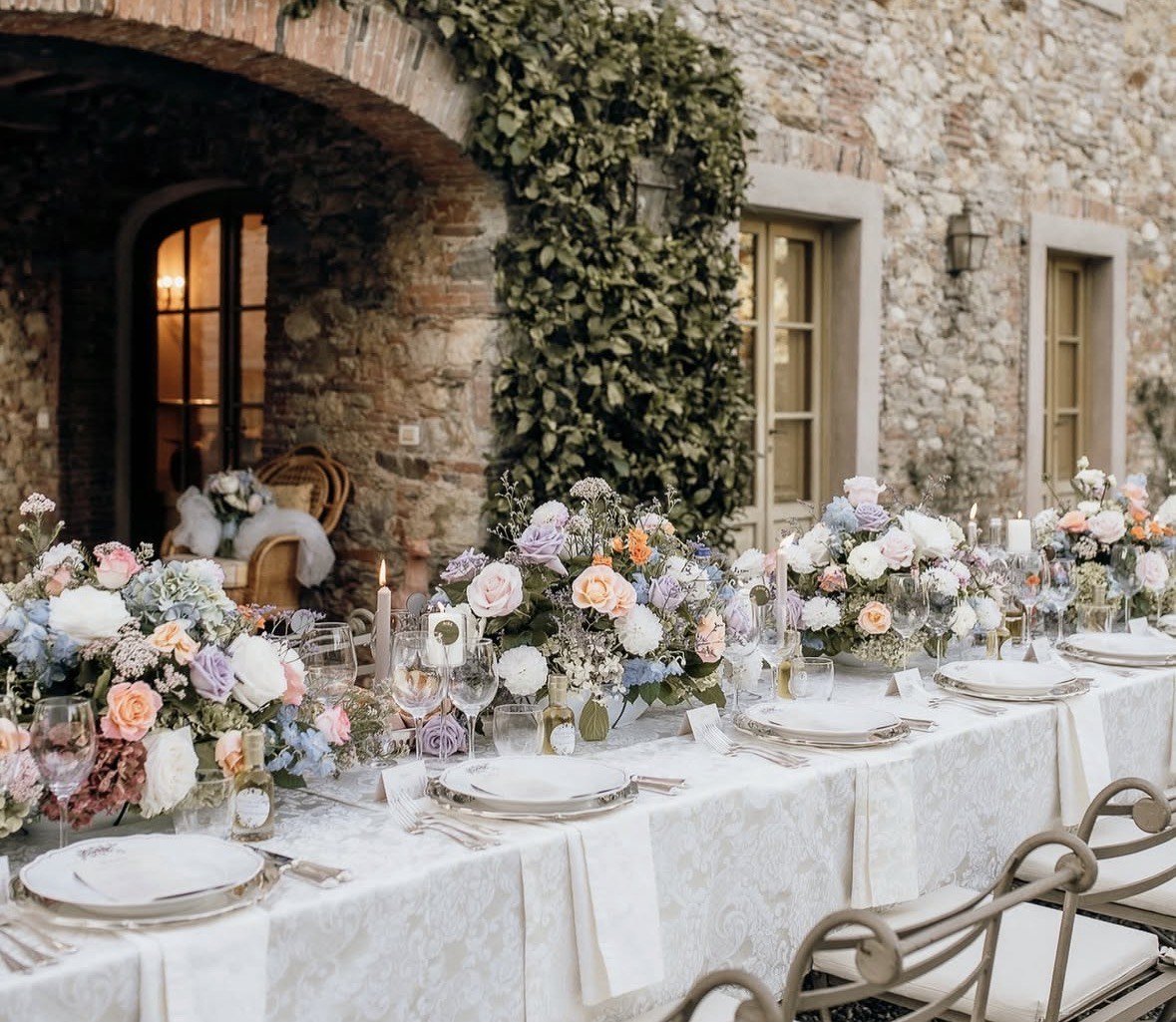 A long reception table at a stone villa set with low arrangements of blush roses, dusty-blue hydrangea and candles