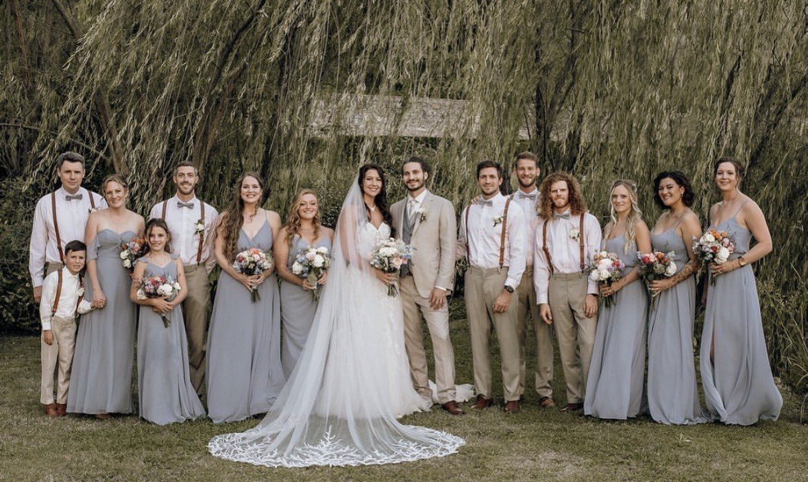 The full wedding party in dusty blue and sand tones lined up beneath a willow tree, each carrying Julie's bouquets