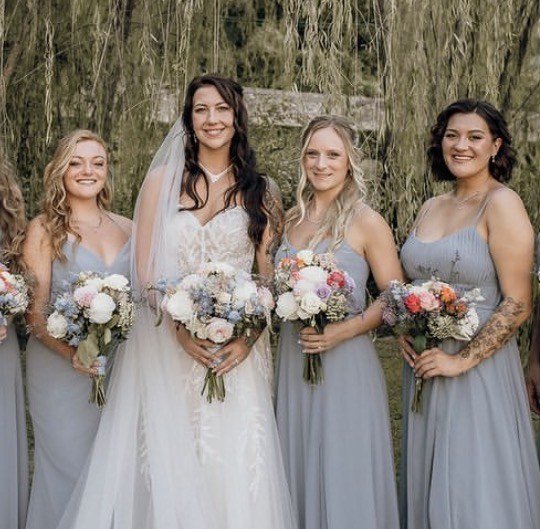 A bride and three bridesmaids in dusty-blue gowns holding matching bouquets of blush roses and blue hydrangea