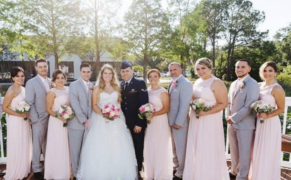 The wedding party in blush gowns and grey suits on a sunlit porch, the bride with a pink and cream rose bouquet