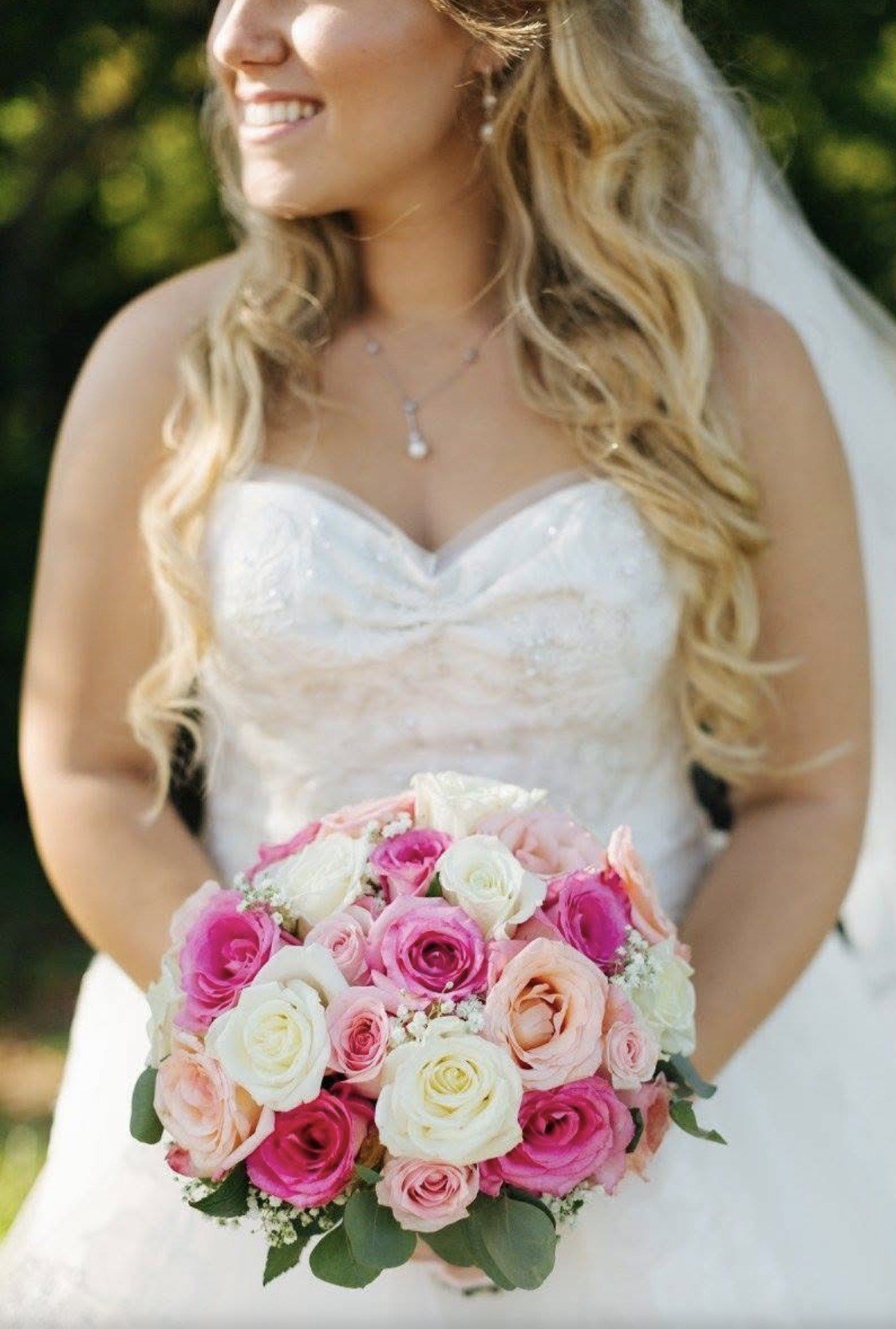 A smiling bride holds a round bouquet of pink, cream and peach roses with eucalyptus