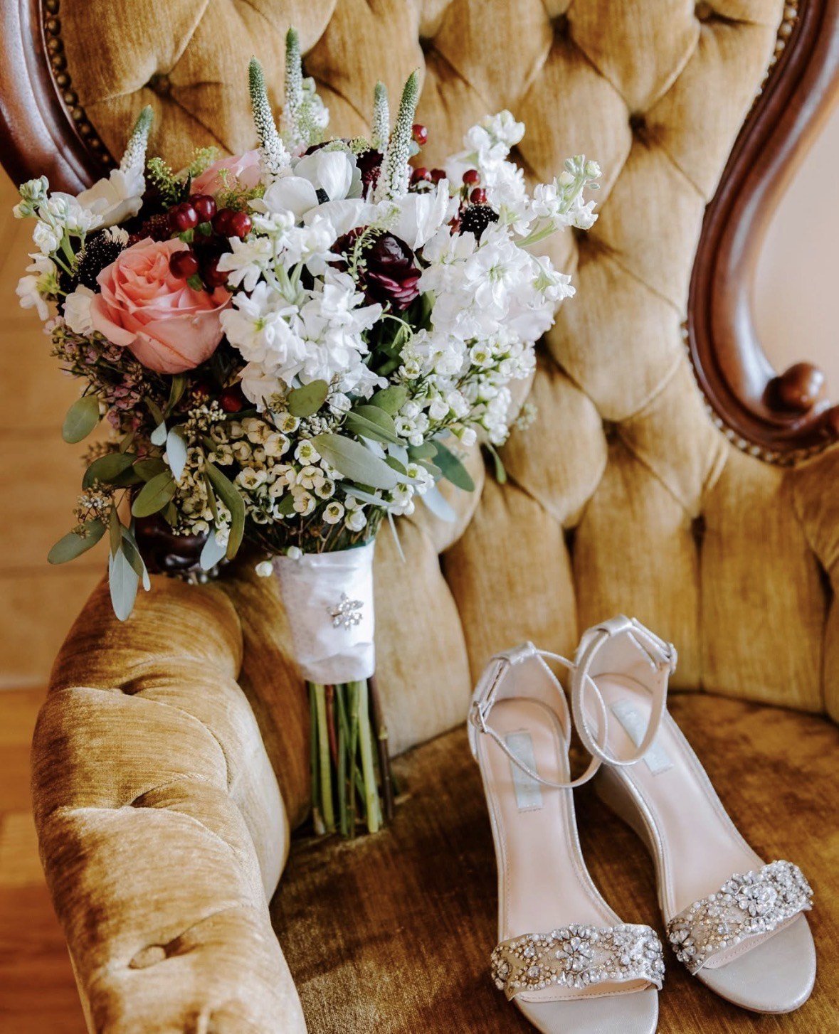 A bouquet of blush roses, burgundy ranunculus and white stock laid on a tufted gold velvet chair beside the bride's shoes