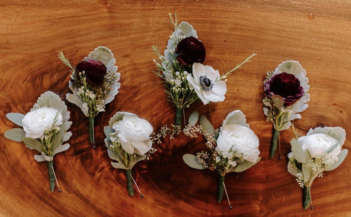 Six boutonnières of white anemone, burgundy ranunculus and eucalyptus laid out on a wooden board