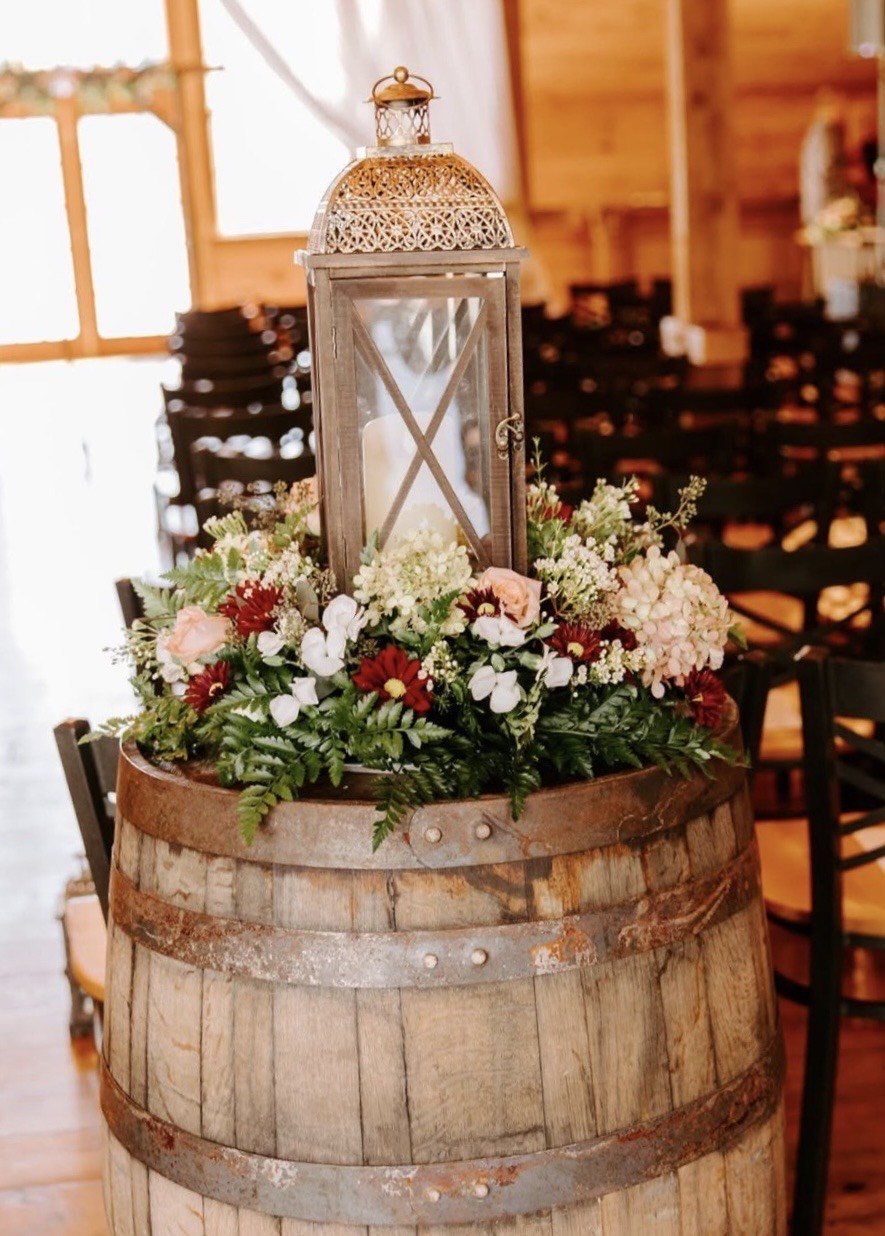A lantern set on a whiskey barrel ringed with blush roses, burgundy dahlias, white blooms and ferns inside the pine chapel