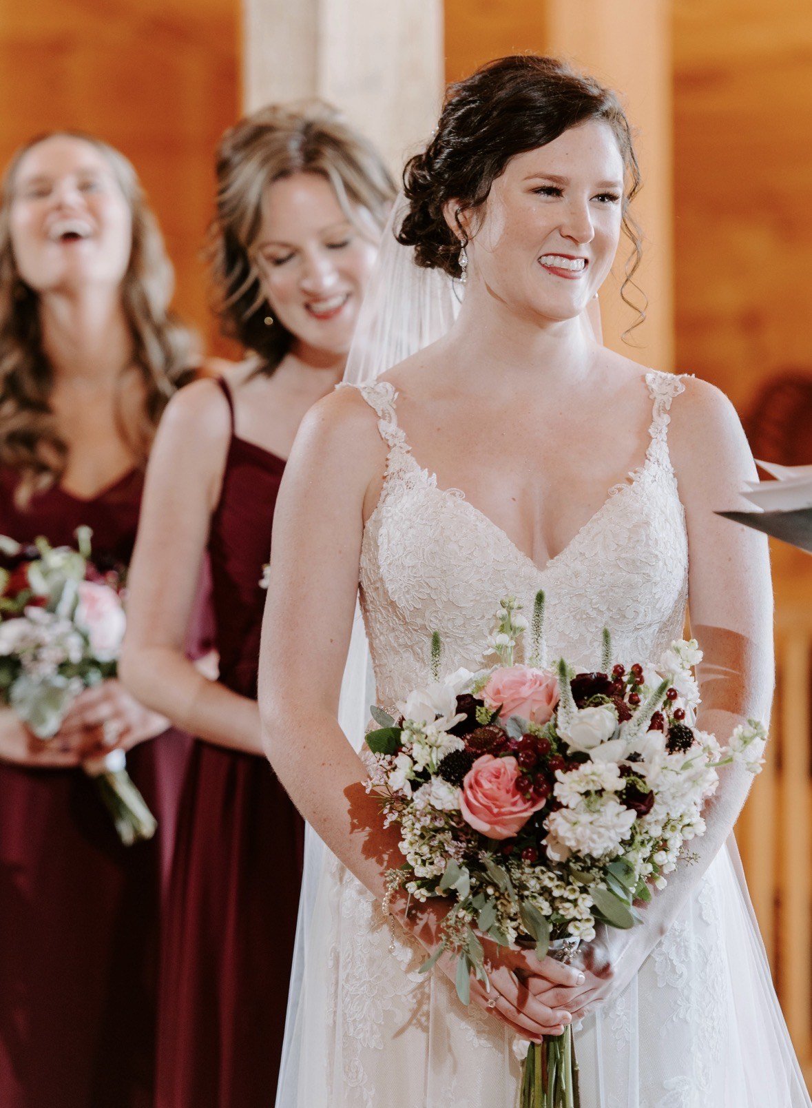 A bride at the altar holding Julie's bouquet of blush garden roses, white stock, veronica and waxflower, bridesmaids in burgundy behind her