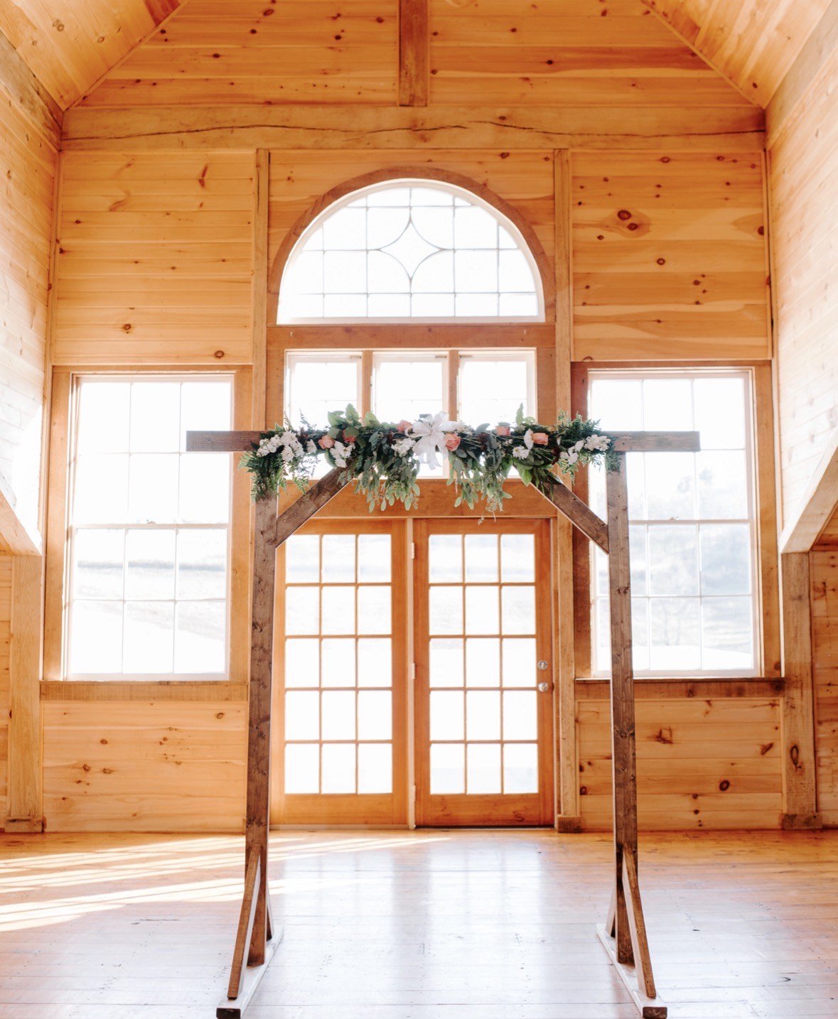 A wooden ceremony arch dressed with Julie's garland of roses, lilies and trailing greenery inside a sunlit pine chapel